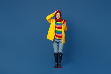 Full body sad young woman in sweater red hat yellow waterproof raincoat use mobile cell phone read forecast isolated on plain dark royal navy blue background Outdoors wet fall weather season concept