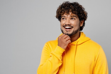Fototapeta na wymiar Young cheerful minded smiling happy Indian man 20s he wear casual yellow hoody look aside on workspace area prop up chin isolated on plain grey background studio portrait. People lifestyle portrait.