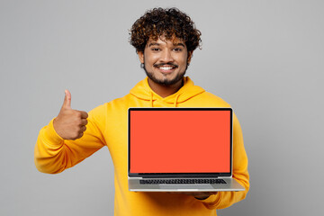 Young smiling happy fun IT Indian man 20s wear casual yellow hoody hold use work on laptop pc computer with blank screen workspace area show thumb up isolated on plain grey background studio portrait.