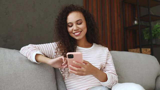 Young happy calm woman wear casual clothes sit on grey sofa couch hold in hand use mobile cell phone in pink case ok sign stay at home hotel flat rest relax spend free spare time in living room indoor