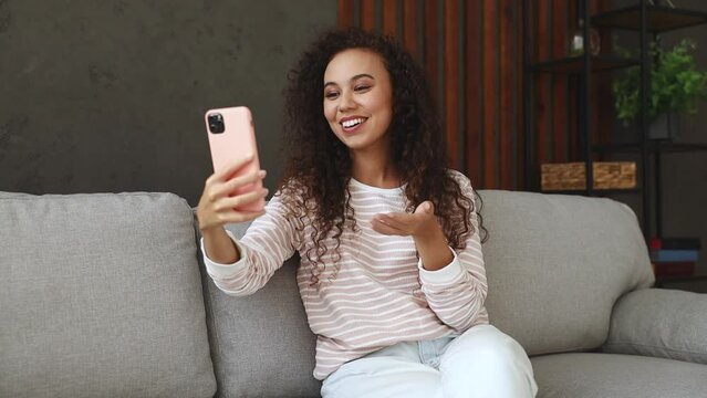Young happy fun woman sits on grey sofa couch stay at home get video call hold use mobile cell phone doing selfie talk greet with hand hotel flat rest relax spend free spare time in living room indoor