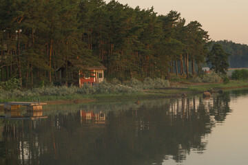 Nagu, Finland - 12.06.2022: A small red house on the shore of the evening bay. The pine tree line...