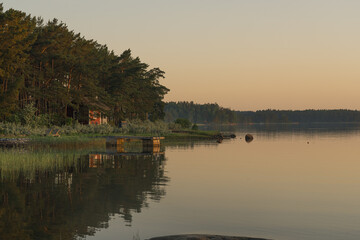 Nagu, Finland - 12.06.2022: A small red house on the shore of the evening bay. The pine tree line...
