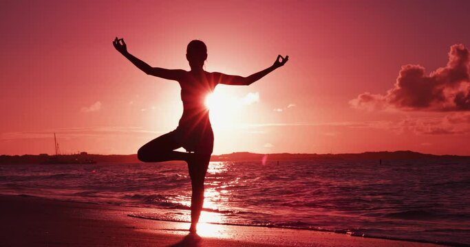 Yoga woman at beach sunset in tree pose meditating outdoors on by ocean sea. Female yoga instructor working out training in serene ocean landscape. Silhouette of woman doing yoga at sunrise