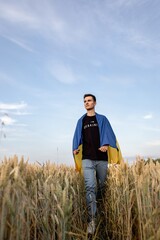 Naklejka na ściany i meble a young guy is walking in a wheat field against the background of a blue sky with textured clouds at sunset with the Ukrainian flag on his shoulders