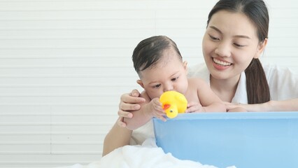 Baby girl holding rubber duck toy to bite and itch gums in blue bathtub while young mother bathing her daughter.