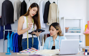 Fototapeta premium Millennial Asian young professional female dressmaker designer seamstress mentor with measuring tape helping teaching advising colleague choosing color pattern chart in tailor studio workshop office