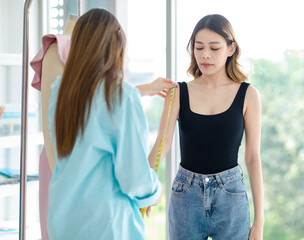 Millennial Asian young professional successful female designer dressmaker seamstress businesswoman using measuring tape measure customer arm length fitting for dress in tailor workshop studio office