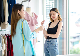 Millennial Asian young professional female dressmaker designer seamstress holding color pattern paper chart helping customer choosing colour for dressing on manikin at tailor workshop studio office