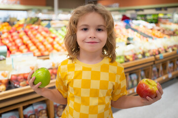 Fototapeta premium Child hold apple fruits at grocery store. Kid is choosing fresh vegetables and fruits in the store. Child buying food in grocery supermarket. Buying in grocery store. Groceries in the supermarket.