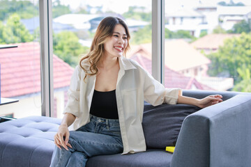 Millennial Asian young beautiful professional female dressmaker designer sitting on cozy sofa at tailor workshop studio.