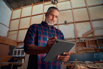 Focused mid adult man using digitized pen on digital tablet near woodpile at woodworking factory