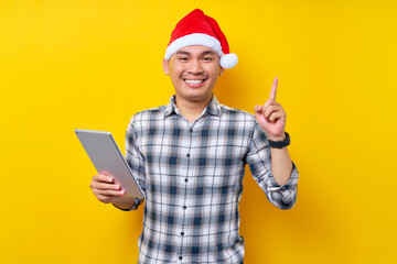 Young handsome smiling Asian man wearing Christmas hat holding tablet, pointing finger up with great new idea on yellow background. Celebration Christmas and Happy New Year 2023 holiday concept