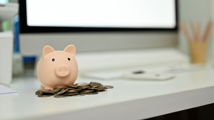 Piggy bank and coins on office desk with blurred background of computer and office supplies.
