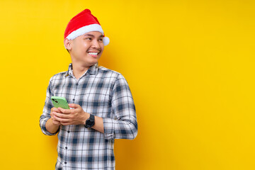 Smiling young Asian man in plaid shirt wearing Christmas hat holding mobile phone and looking aside at the workspace area on yellow background. Christmas and Happy New Year 2023 holiday concept