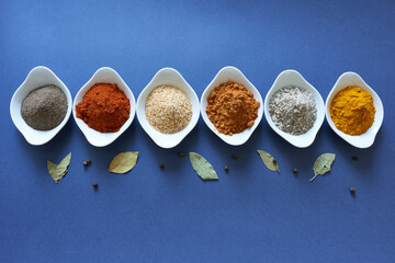 A variety of spices in white bowls on a blue background with dried laurel leaves. 