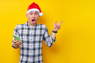 Surprised young Asian man in plaid shirt wearing Christmas hat holding mobile phone and pointing index finger aside isolated on yellow background. Christmas and Happy New Year 2023 holiday concept