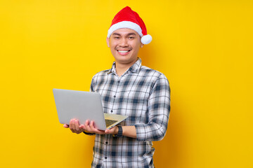 Smiling young Asian man in plaid shirt wearing Christmas hat posing holding laptop pc computer, looking confident at camera on yellow background. Christmas and Happy New Year 2023 holiday concept
