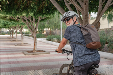 Smiling active cyclist senior man in urban park wearing helmet and backpack running with electric bicycle.  Concept of healthy lifestyle and sustainable mobility