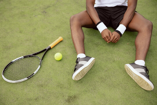 Close-up of the hands and legs of African-looking tennis player sitting on the grass of tennis court. Next to him lies tennis racket and ball. Guy is wearing white T-shirt, black shorts and sneakers.