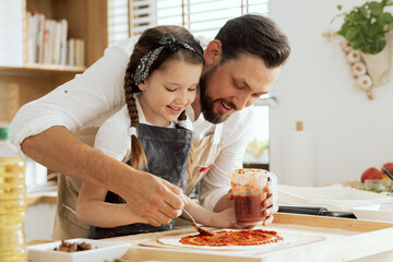 Happy mother's day handsome father with young daughter applying ketchup on homemade dough for pizza...