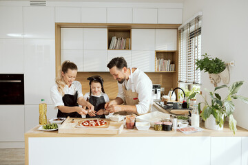 Happy young mother and father with adorable daughter standing at modern kitchen dressing domestic...