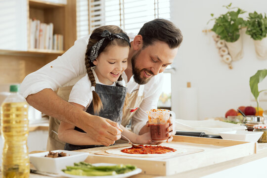 Adorable young daughter with lovelu father in aprons applying kutchup on fresh kneaded homemade dough for pizza. - Powered by Adobe