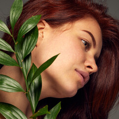 Portrait of tender red-haired woman posing with leaves isolated over dark grey background. Concept of skin care, cosmetology, natural hair and skin cosmetics