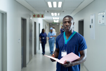 Portrait of african american male healthcare worker with file in busy hospital corridor, copy space