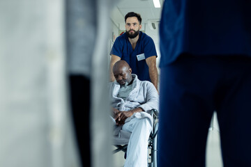 Diverse male healthcare worker pushing african american male patient in wheelchair in hospital