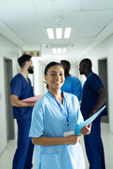 Fototapeta premium Vertical portrait of smiling biracial female healthcare worker in busy hospital corridor, copy space
