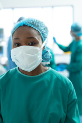 Fototapeta na wymiar Vertical portrait of african american female surgeon wearing face mask in operating theatre