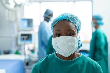 Fototapeta na wymiar Portrait of african american female surgeon in face mask in operating theatre, with copy space