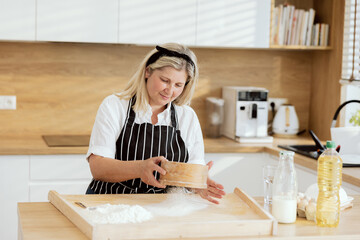Elderly woman sieving flour on wooden surface wearing apron standing at table in modern kitche cooking baking cookies caked pizza pasta.