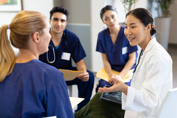 Smiling asian female doctor with tablet sitting in discussion with diverse colleagues at hospital