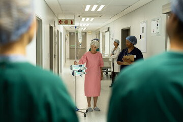Senior caucasian female patient with iv pole on wheels walking in busy hospital corridor