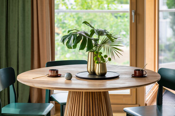 Creative composition of dinning room interior with wooden round table, gold vase with dried flowers, black trace, gray chairs, curtain, cups, window and personal accessories. Home decor. Template.