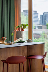 Contemporary design of kitchen interior with wooden kitchen island, modern red barstools, vase with rhubarb, zucchini, strawberries, green curtain and personal accessories. Home decor. Template.