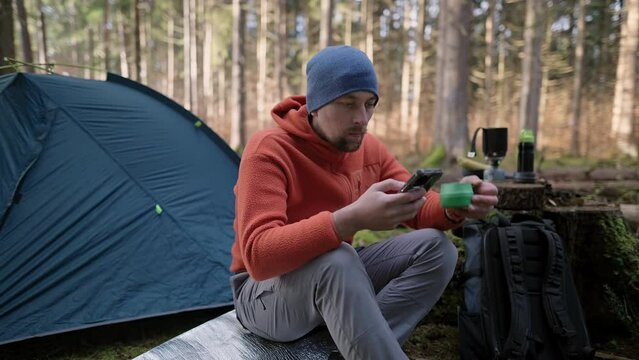 Male hiker drinking tea and surfing Internet on smartphone in tent camp during hiking in fall in woods. Backpacker makes route in map in phone and drinks hot drink from cup in background of tent. 
