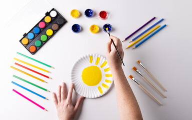 Overhead of hands painting with yellow paint on paper plate, with art materials on table top