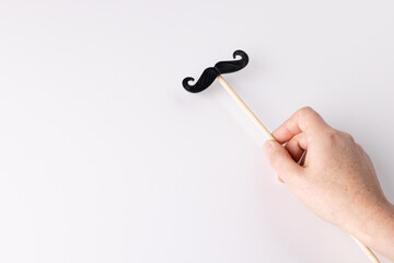 Hand holding black false moustache on stick, on white background with copy space