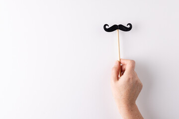 Hand holding black false moustache on stick, on white background with copy space