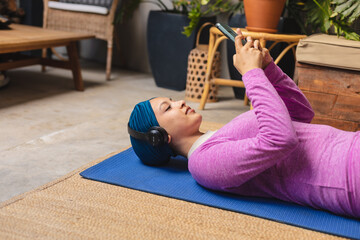 Happy biracial woman wearing hijab, laying on yoga mat and using smartphone in living room