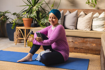 Happy biracial woman wearing hijab, sitting on yoga mat and using smartphone in living room