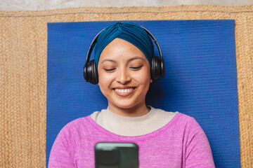Happy biracial woman wearing hijab, laying on yoga mat and using smartphone in living room