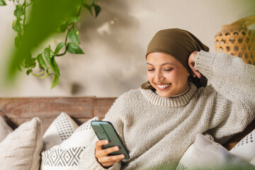 Happy biracial woman wearing hijab, sitting on sofa in living room, using smartphone