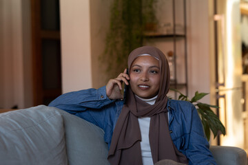 Happy biracial woman wearing hijab, sitting on sofa in living room, talking on smartphone