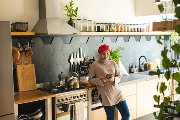 Happy biracial woman wearing hijab, standing in kitchen and using smartphone