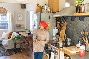 Happy biracial woman wearing hijab, standing in kitchen and using smartphone