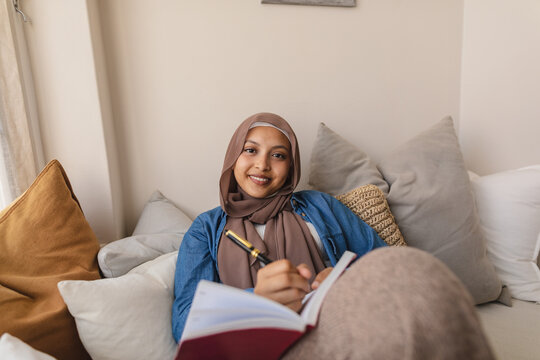 Portrait of happy biracial woman wearing hijab, sitting on sofa and taking notes